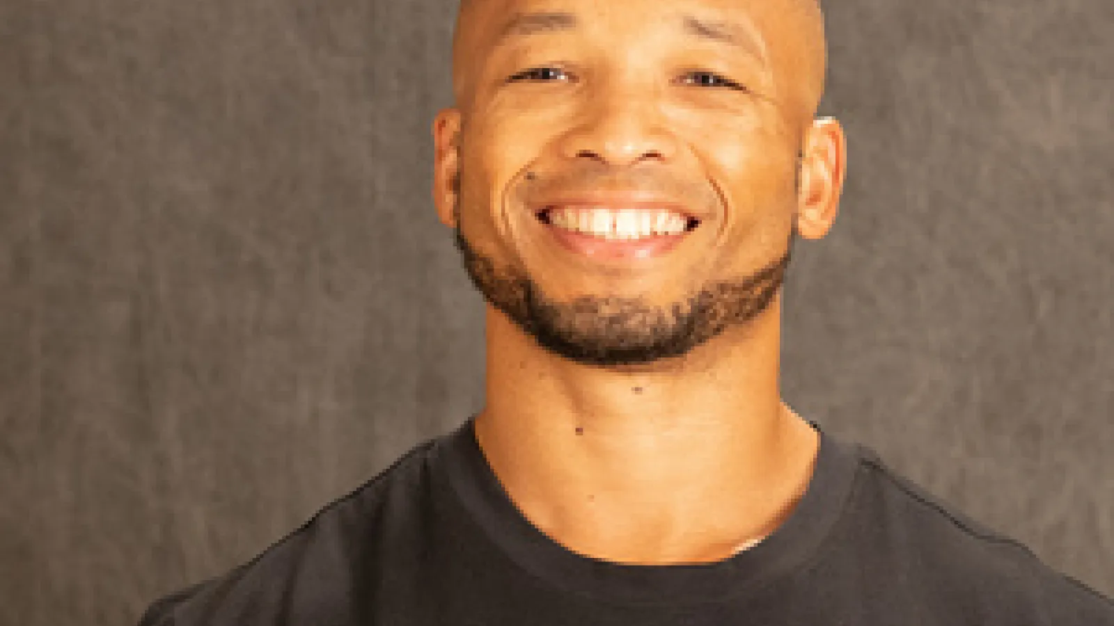 OPEEP Advisory Council member and Lecturer in Department of Sociology at OSU smiles while standing for a professional headshot against a dark gray background while wearing a black t-shirt with a white-and-red OSU logo and a white Nike swoosh on the front.