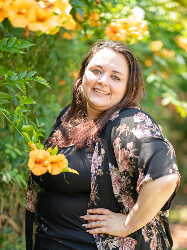 Woman-presenting person standing in front of green leaves and yellow flowers smiling. 