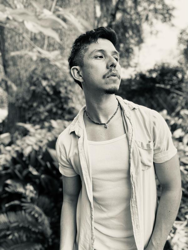 Black and white photo of Irvin Gonzalez wearing a white shirt against a background of trees and foliage.