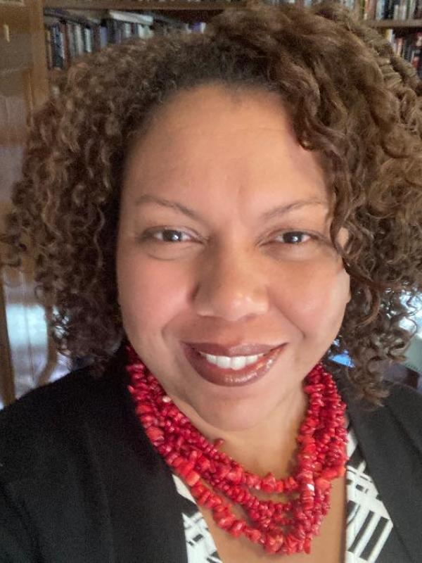 Woman with brown skin and brown curly hair smiles wearing a red necklace and black-and-white shirt.
