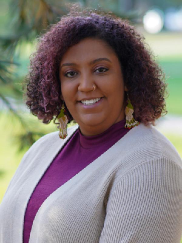 Smiling woman with brown skin, curly purple hair, and long dangly earrings.